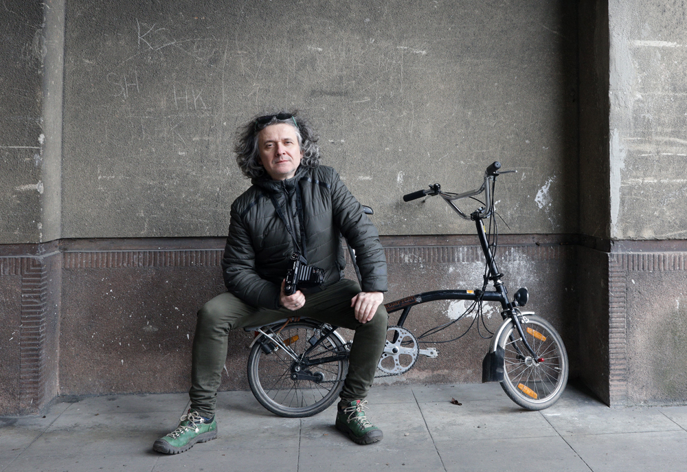 Maciej Rawluk, mężczyzna w średnim wieku o szpakowatych włosach do ramion siedzi na składanym rowerze z aparatem w ręku