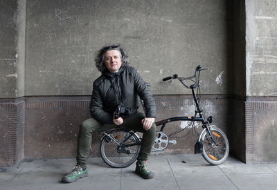Maciej Rawluk, a middle-aged man with shoulder-length gray hair, sitting on a foldable bike with a camera in his hand 