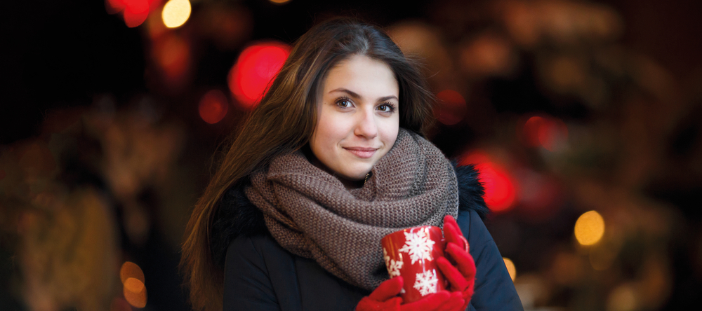 a smiling woman with long hair wearing a jacket, brown scarf and red gloves, holding a red mug with white snowflakes, Christmas lights in the background