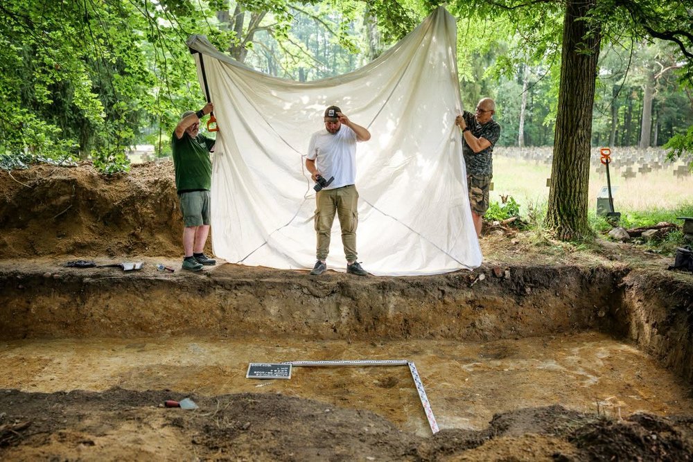 Fotograf stojący nad wykopem za nim biała płachta osłaniająca wykop przed niepożądanym światłem 