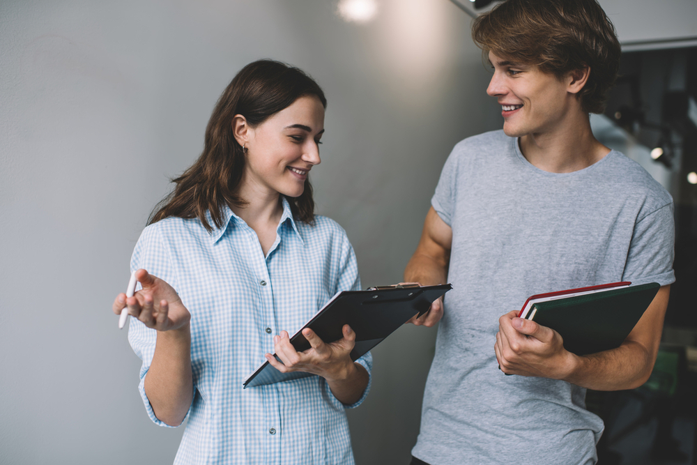 Two smiling students are standing next to each other — one is holding notebooks, the other a clipboard.