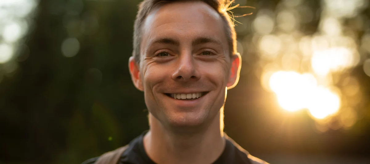 kolorowe zdjęcie przedstawia szeroko uśmiechniętego młodego mężczyznę, za którego plecami przebijają się promienie zachodzącego słońca delikatnie prześwietlające liście otaczających naszego bohatera drzew